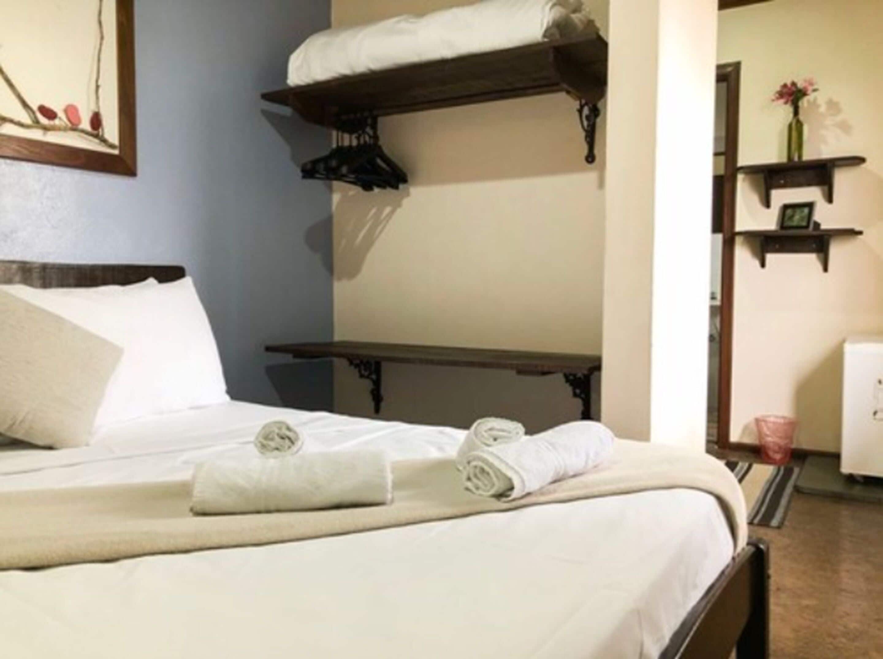A neatly made bed with white linens and rolled towels, a wooden nightstand, and a wall-mounted shelf with a vase of flowers.