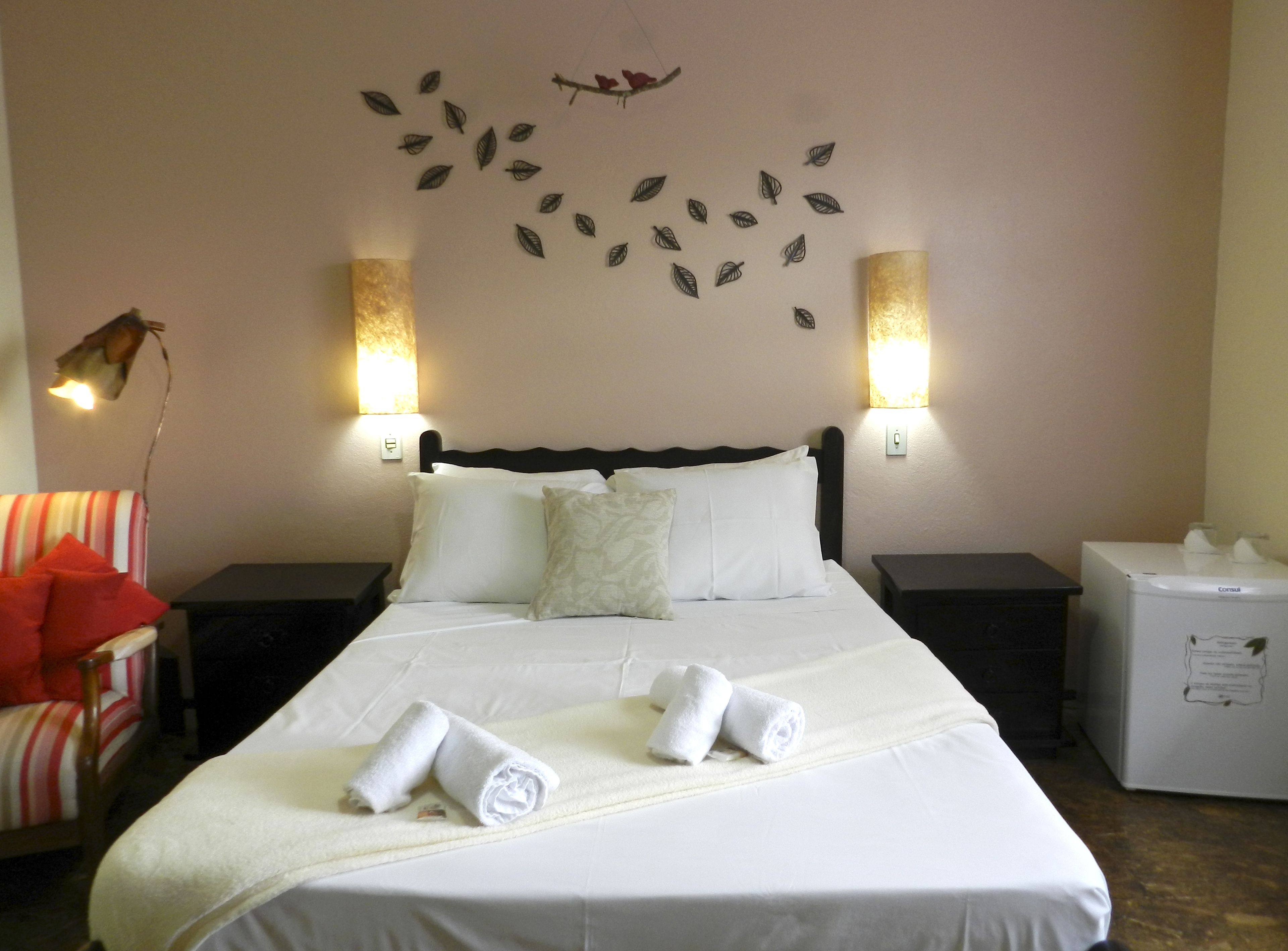 A neatly made bed with white linens and two rolled towels, a wall-mounted light fixture, and a small decorative wall piece.