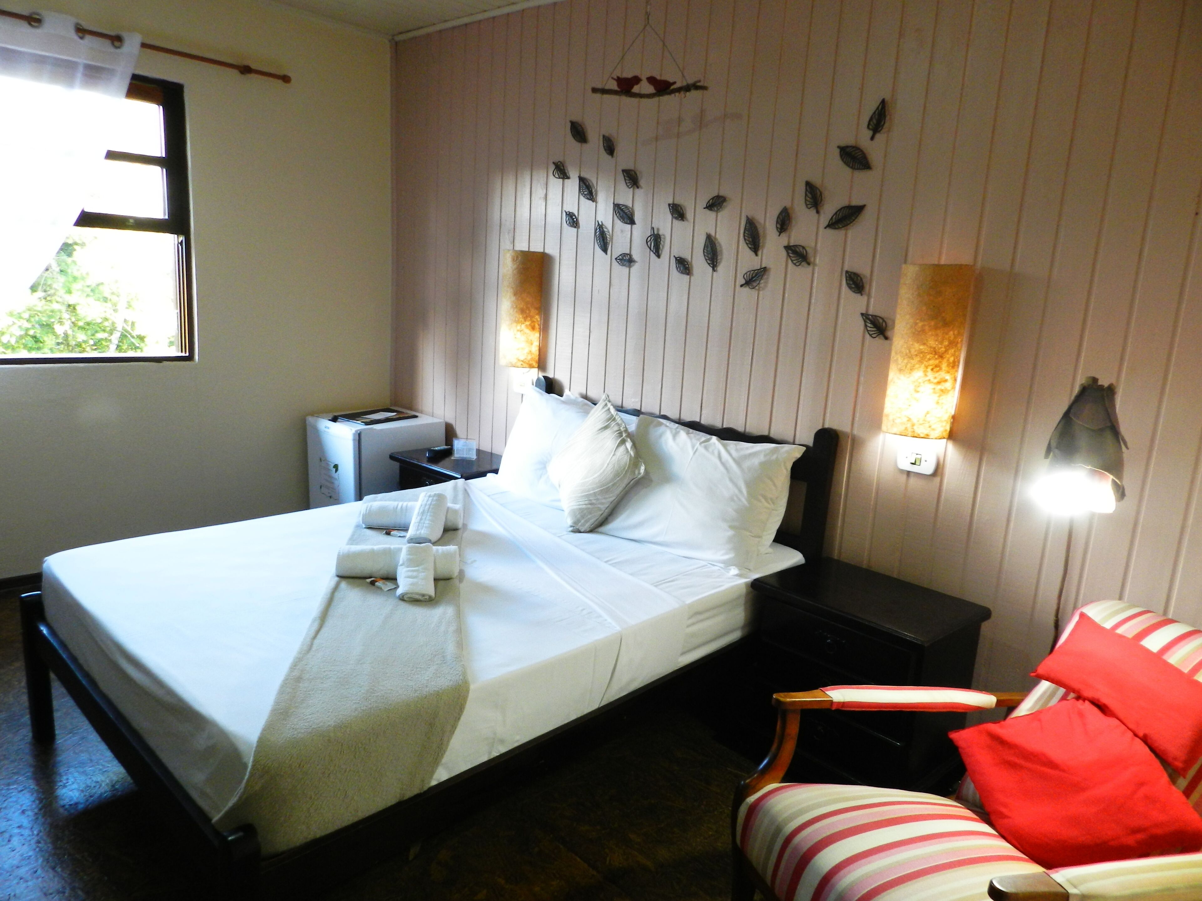 Una cama bien tendida con sábanas blancas, una mesita de noche con una lámpara, una silla a rayas y una pared con diseños de hojas decorativas.