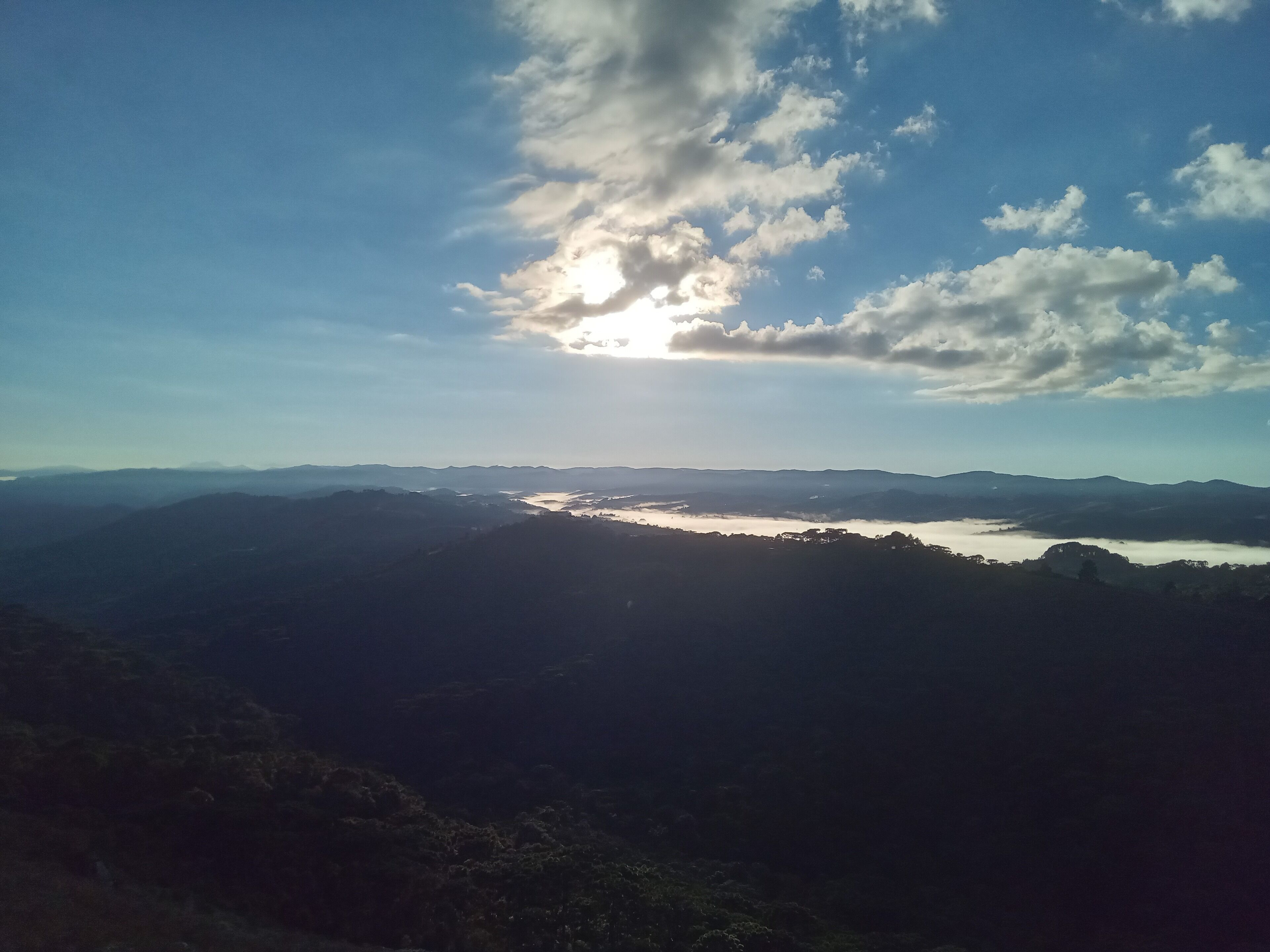 Une vue panoramique d’une vallée traversée par une rivière, entourée de collines et surplombée d’un ciel partiellement nuageux.