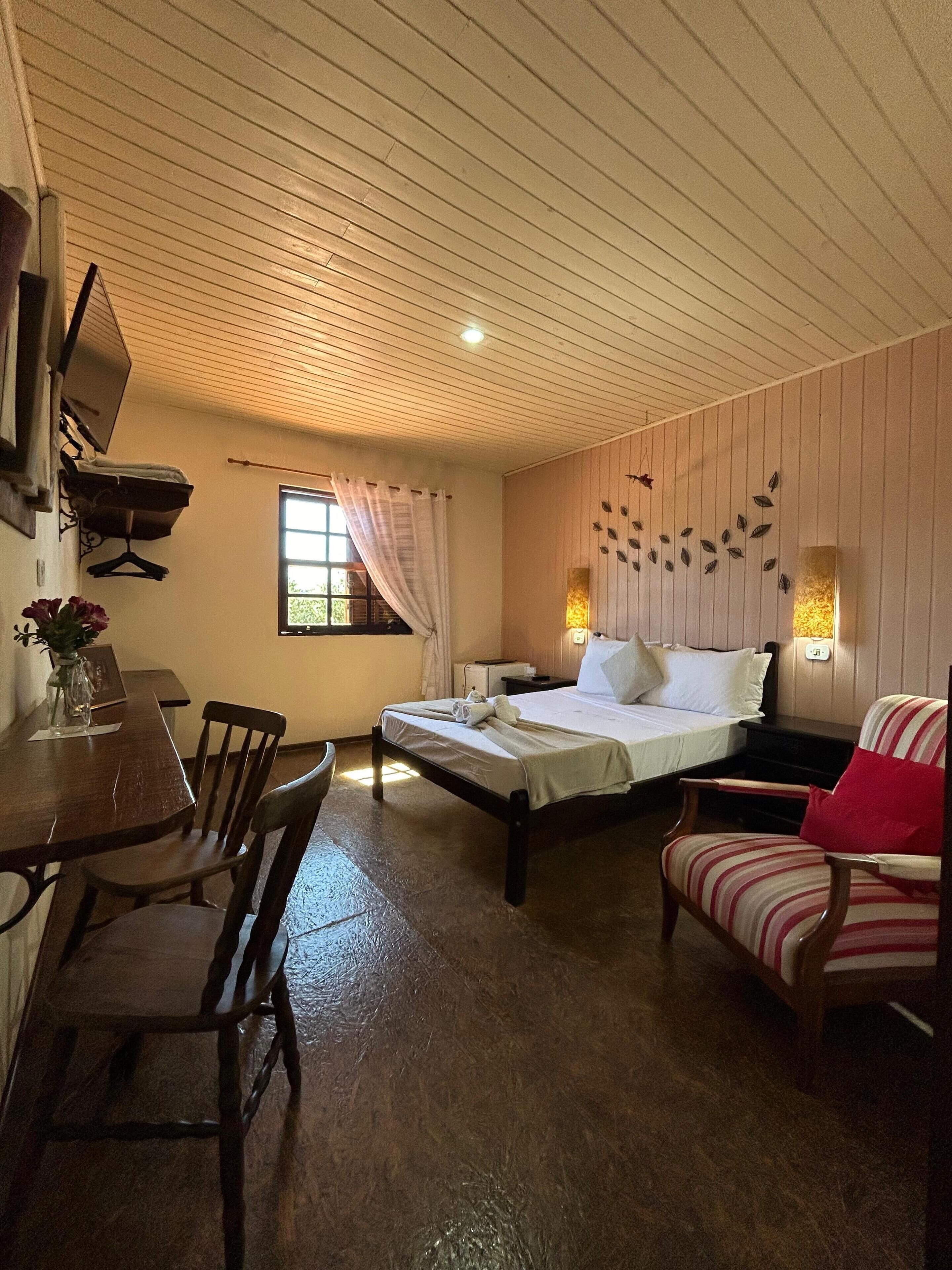 A cozy room with a bed, a desk, and a chair. There is a window with curtains, a wall-mounted TV, and a decorative wall piece.