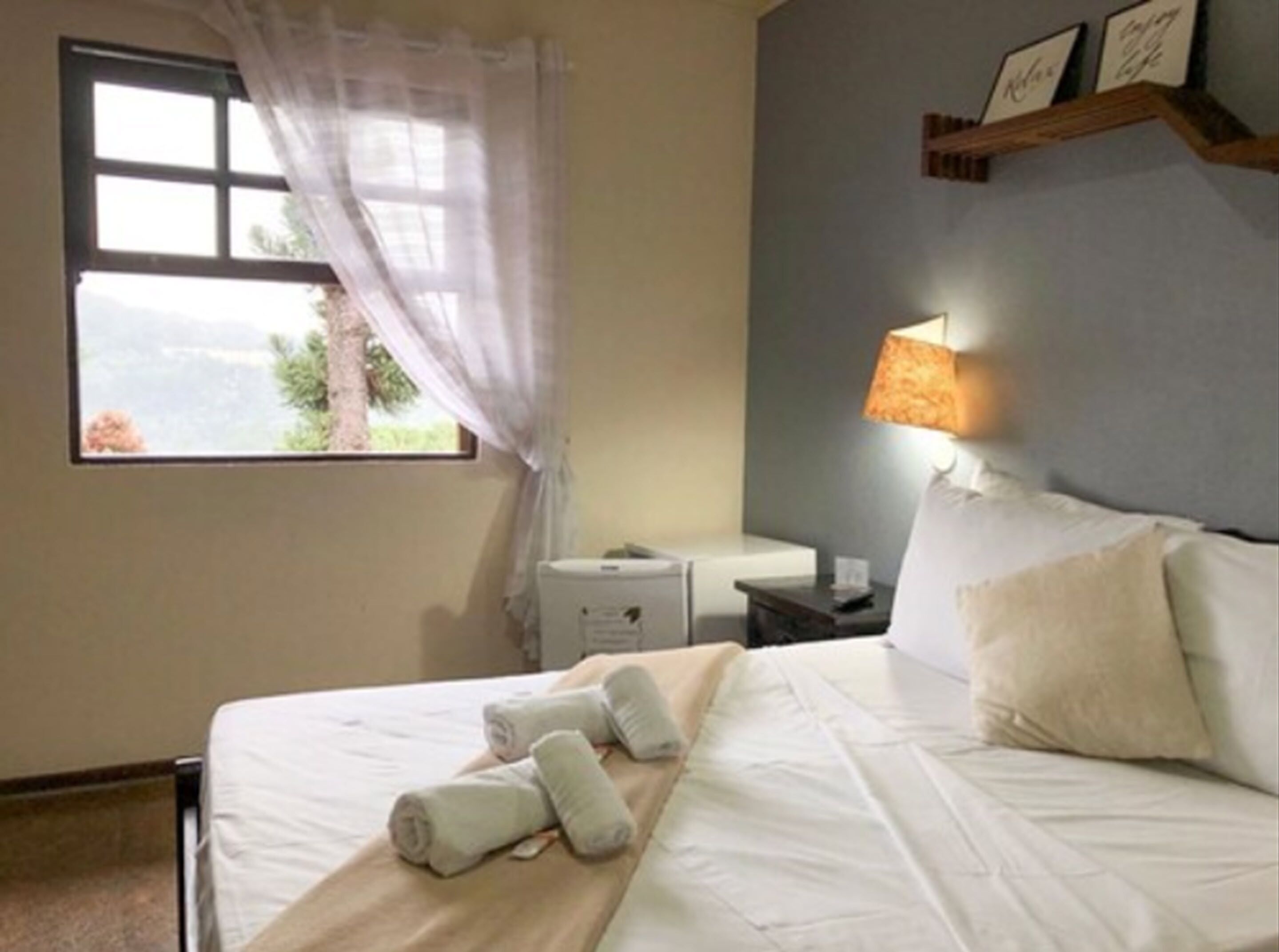 Una habitación de hotel con cama, mesita de noche, lámpara y ventana con vistas a árboles.