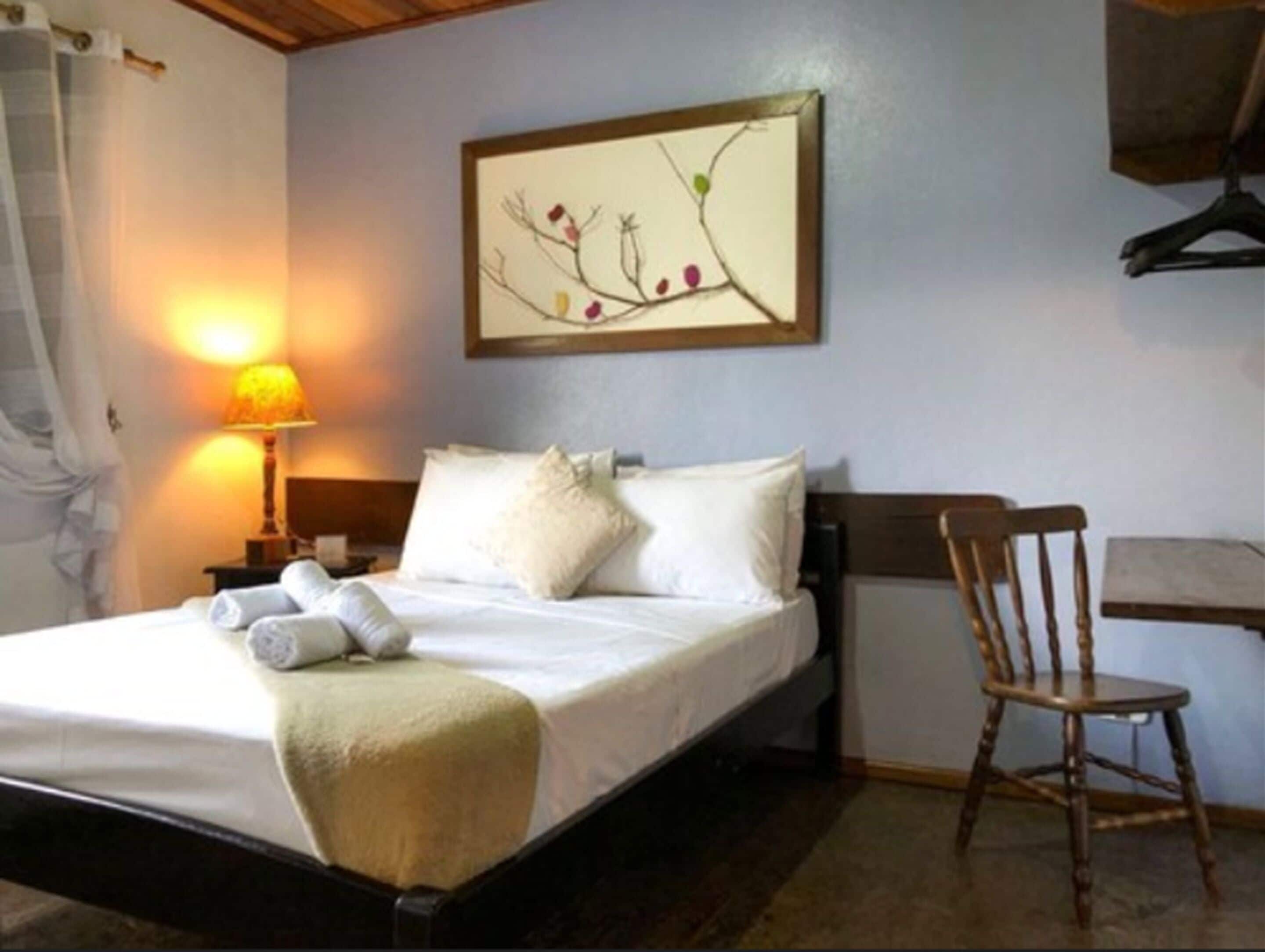 A neatly made bed with white linens and a beige throw blanket, a wooden chair, a nightstand with a lamp, and a wall-mounted picture of a tree branch.