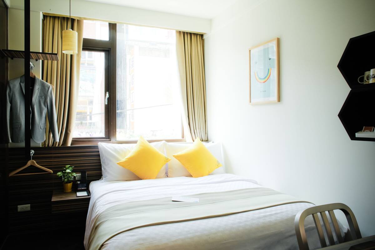 Habitación de hotel con cama, escritorio con lámpara, perchero y ventana con cortinas.