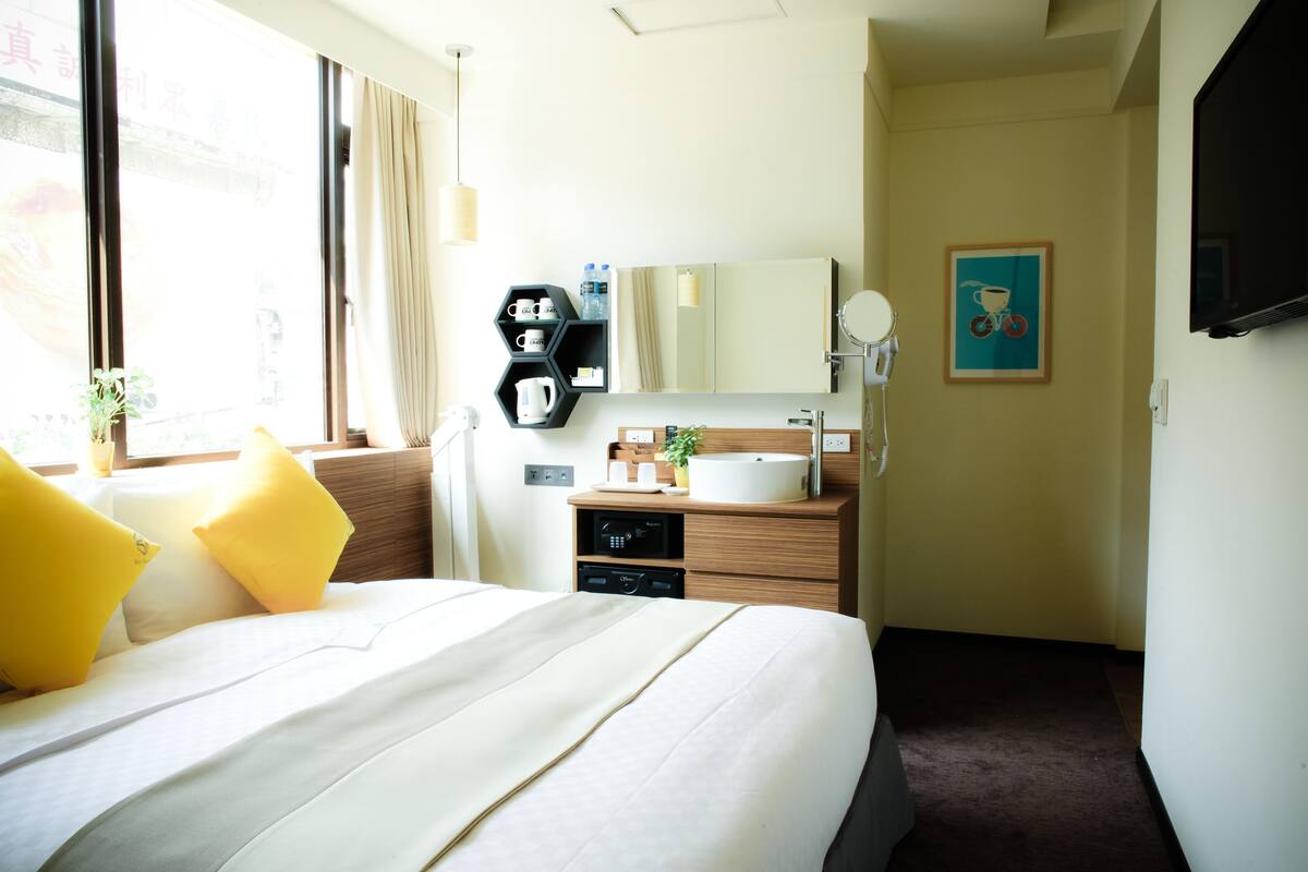 Habitación de hotel con una cama grande, una mesita de noche con cafetera, un espejo y un televisor montado en la pared.