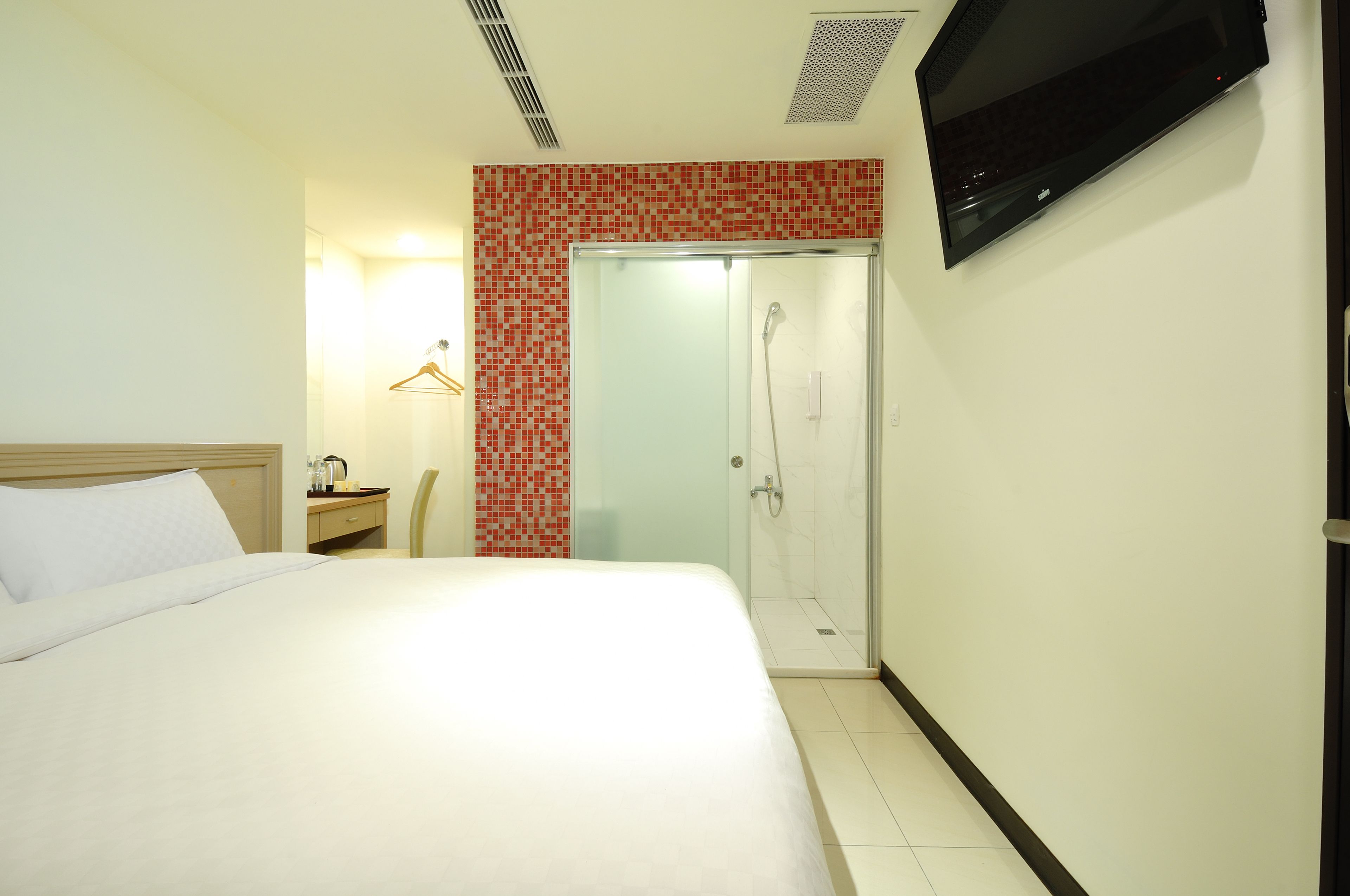 A hotel room with a bed, a TV mounted on the wall, a shower, and a small table with a kettle.