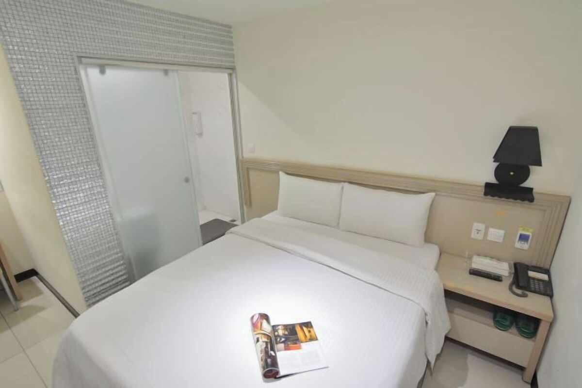 A hotel room with a bed, a nightstand with a lamp, a phone, and a small table with a magazine.