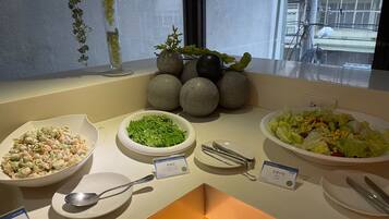 A buffet table with various dishes, including a salad, a bowl of green vegetables, and a bowl of pasta salad. There are also plates and utensils on the table.