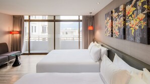 Habitació d'hotel amb dos llits, un sofà, una taula de vidre i vista a la ciutat a través de grans finestres.