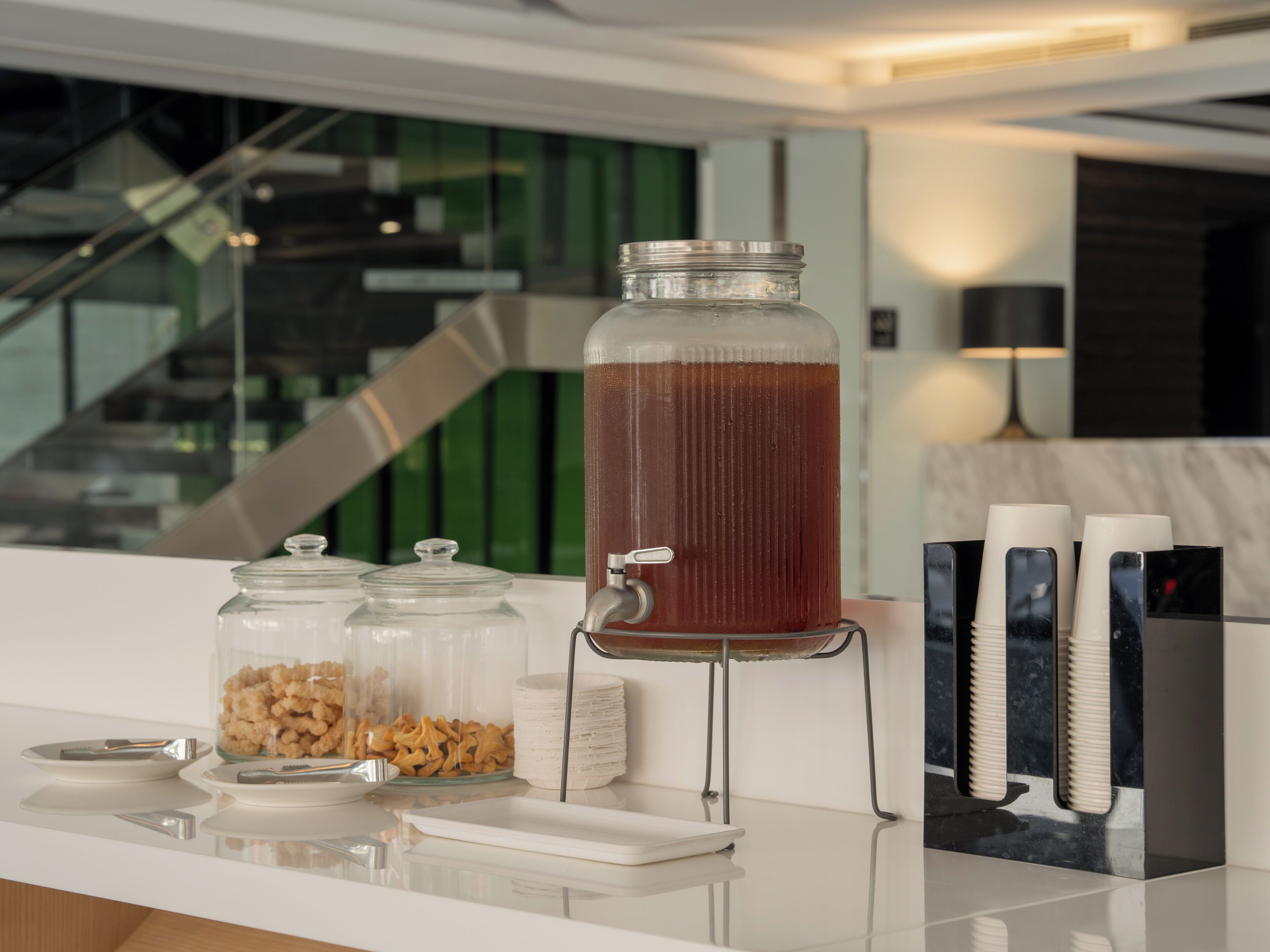 Un dispensador de bebidas con un frasco grande lleno de un líquido marrón, una pila de platos blancos y un recipiente con aperitivos sobre una encimera.