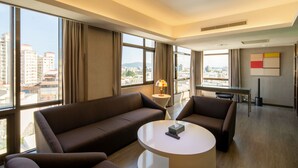 Habitació d'hotel moderna amb un gran finestral, un sofà, una cadira, una taula de centre rodona i una taula petita amb una làmpada.