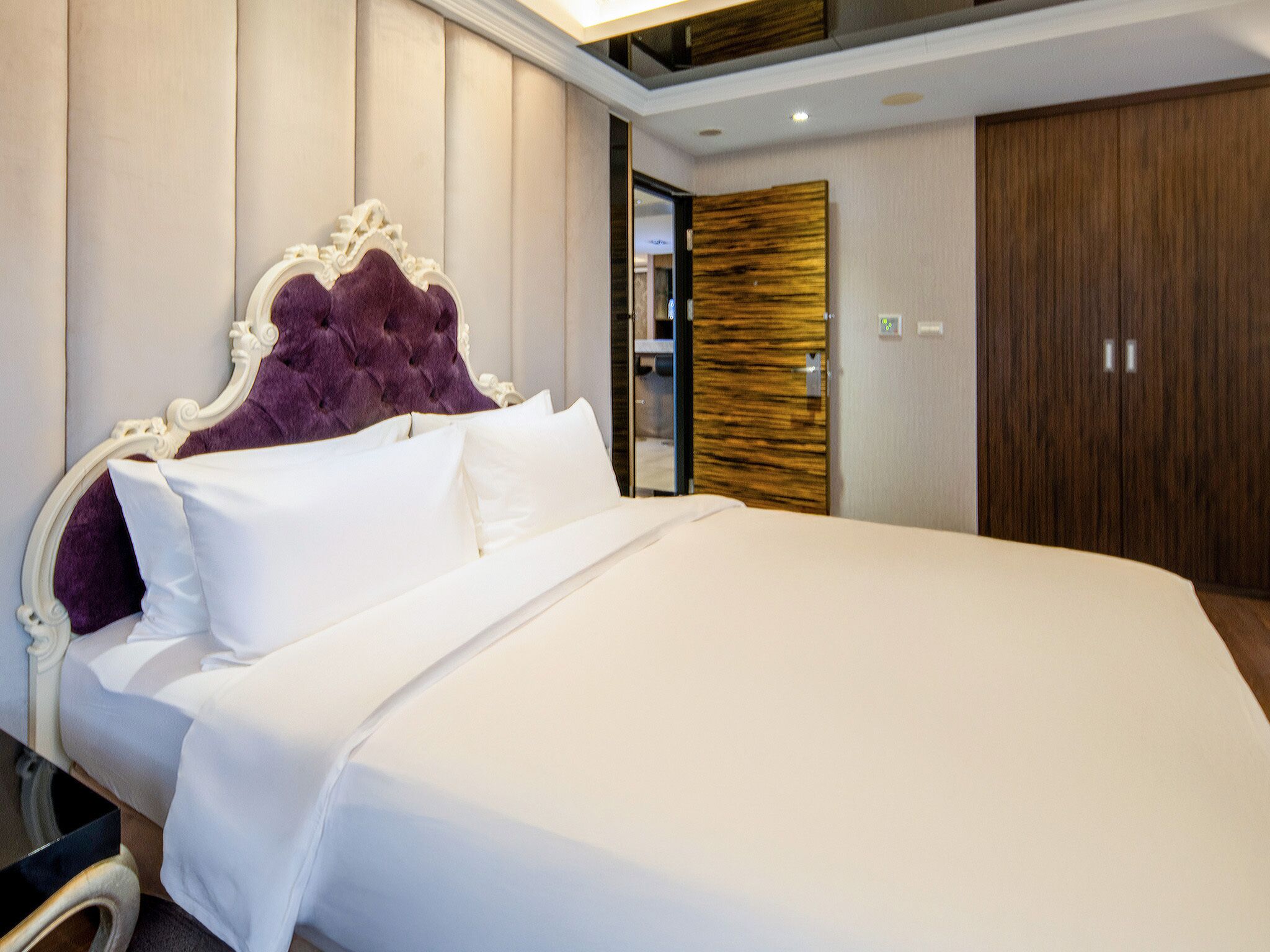 A large bed with white linens and a purple tufted headboard, a wooden wardrobe, and a modern ceiling design.