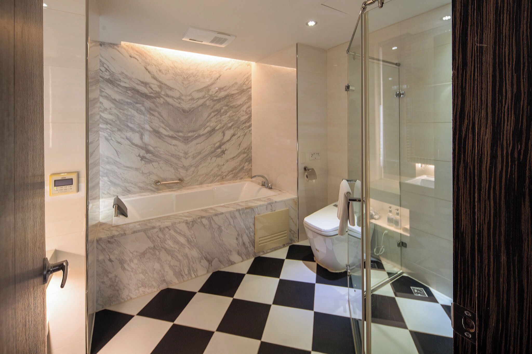A modern bathroom with a marble wall, a black and white checkered floor, a glass shower enclosure, and a white toilet.