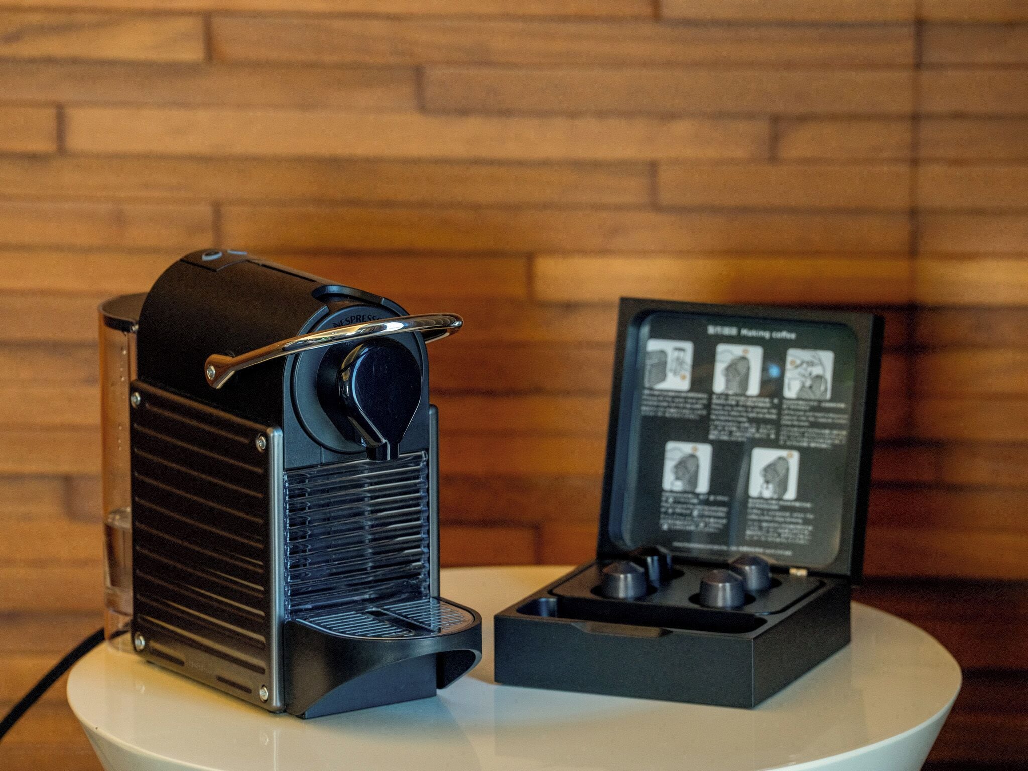 Nespresso-kahvinkeitin ja sen mukana toimitettu käyttöohje pöydällä, taustalla tiiliseinä.