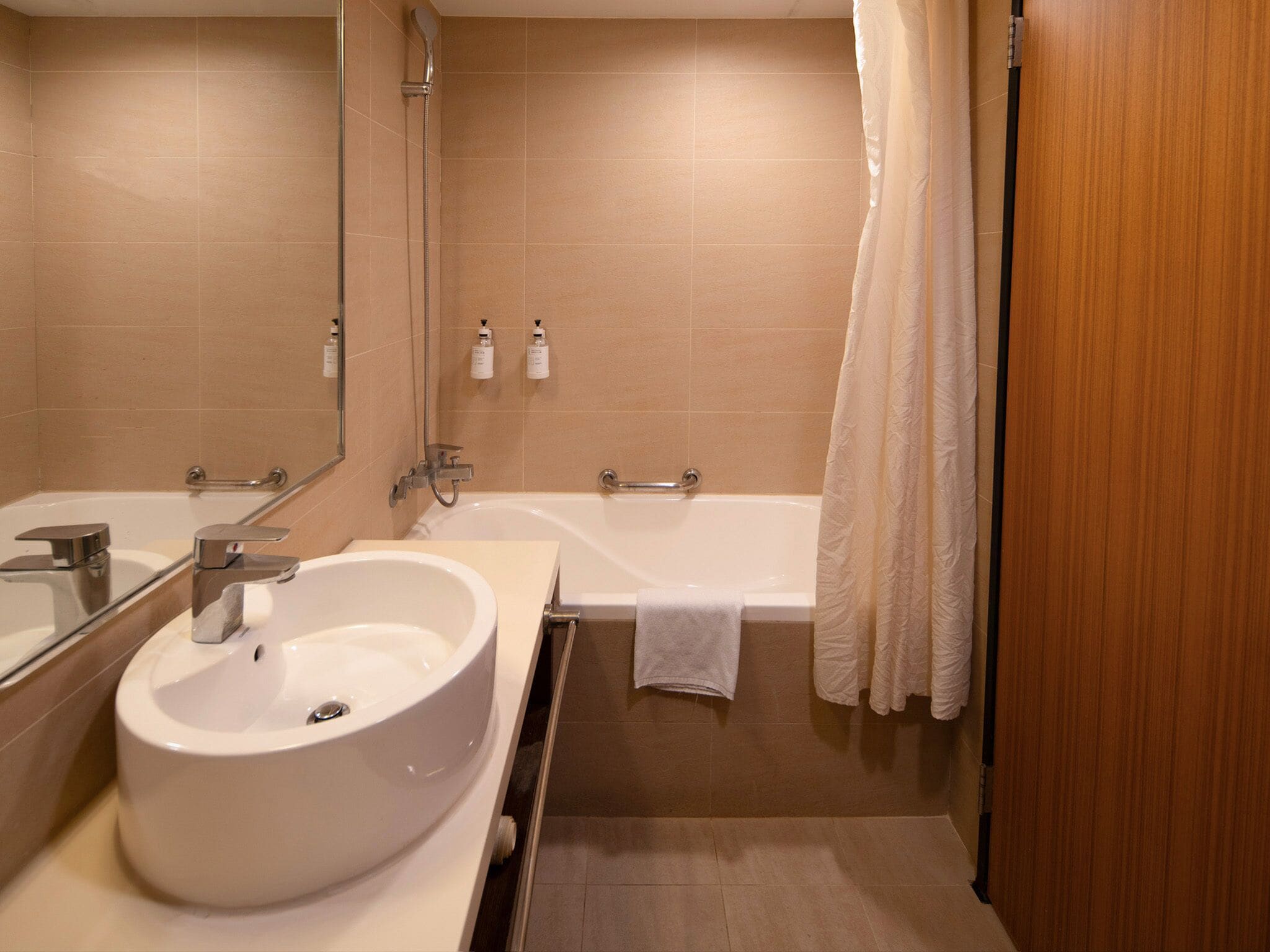 A modern bathroom with a large white sink, a bathtub, and a shower. There are two mirrors, one above the sink and one above the bathtub. The walls are tiled in a light beige color, and there is a wooden door visible on the right side.