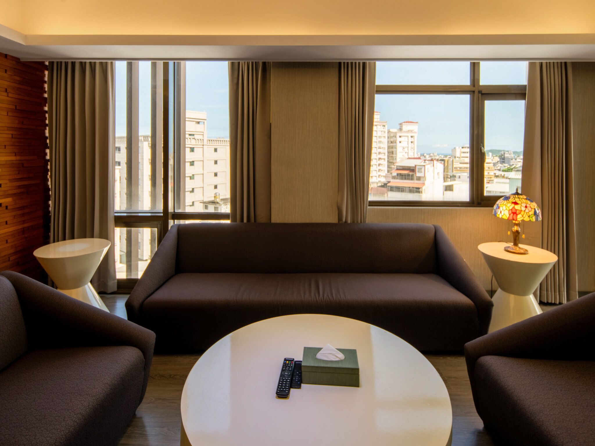 Habitación de hotel con un ventanal grande, un sofá, dos sillones, una mesa de centro y una lámpara.