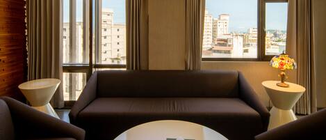 Habitación de hotel con un ventanal grande, un sofá, dos sillones, una mesa de centro y una lámpara.