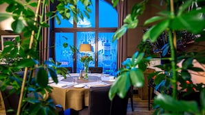A dining area with a table set for four, a vase with flowers, and a view of a cityscape through large windows.