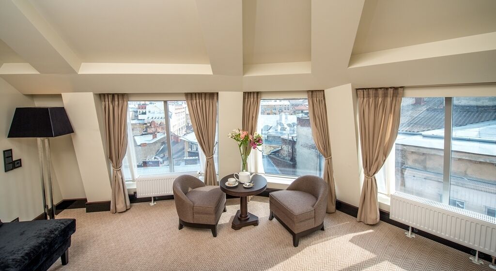 A hotel room with a large window, two armchairs, a small table with a vase, and a lamp.