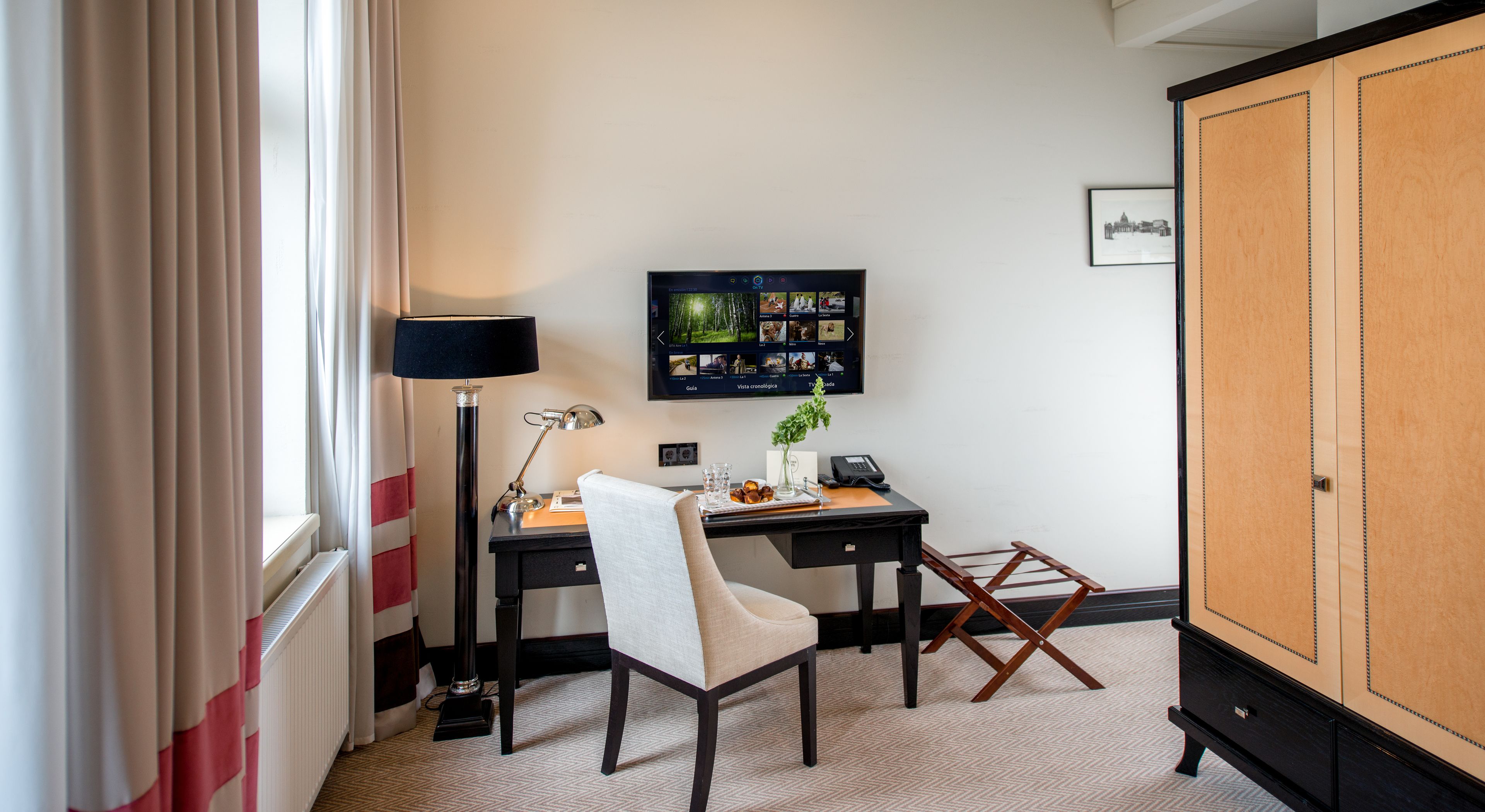 Habitación de hotel con escritorio, silla, lámpara, televisor y armario.