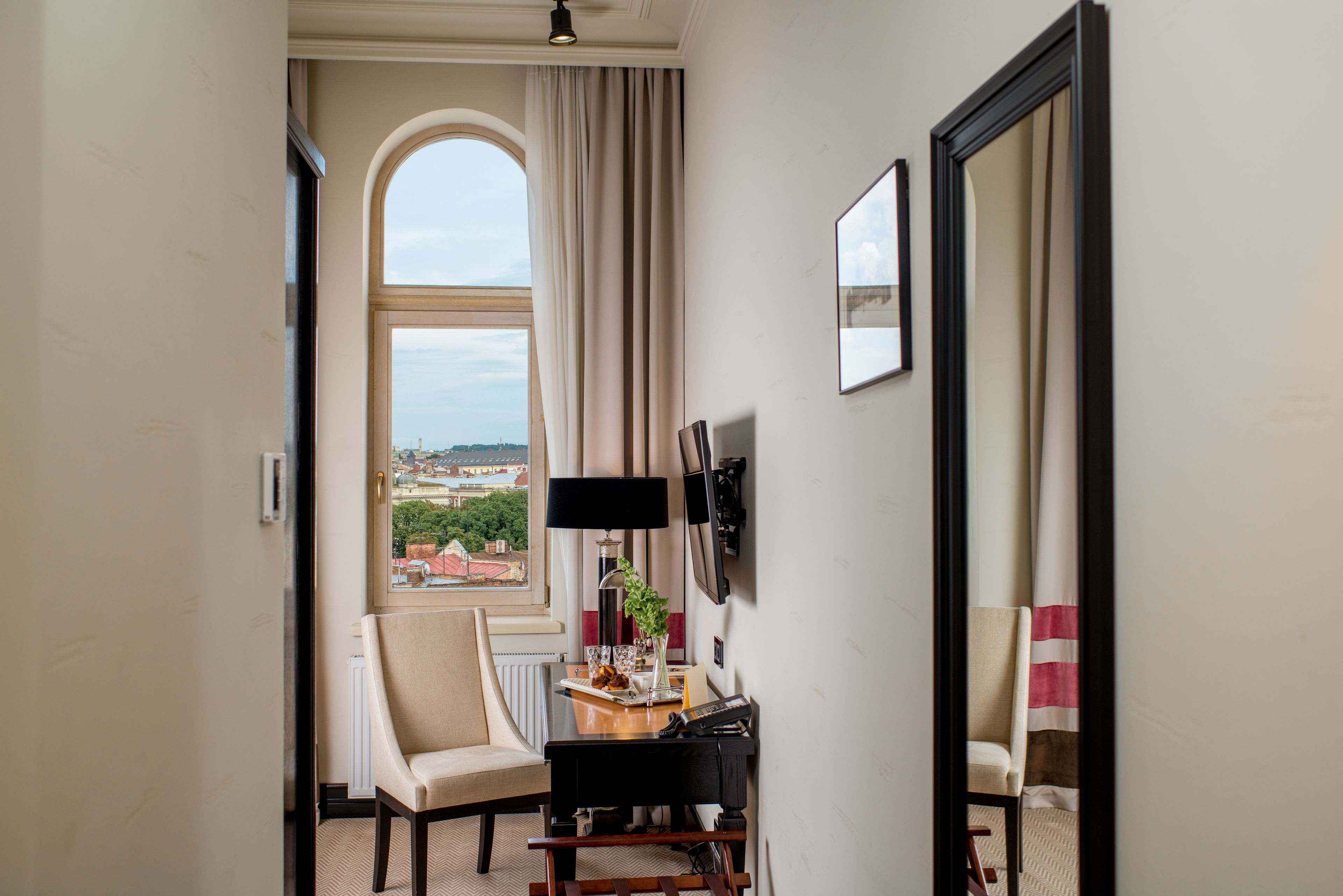 Habitación de hotel con un gran ventanal arqueado, un escritorio con lámpara, una silla y vistas a la ciudad.