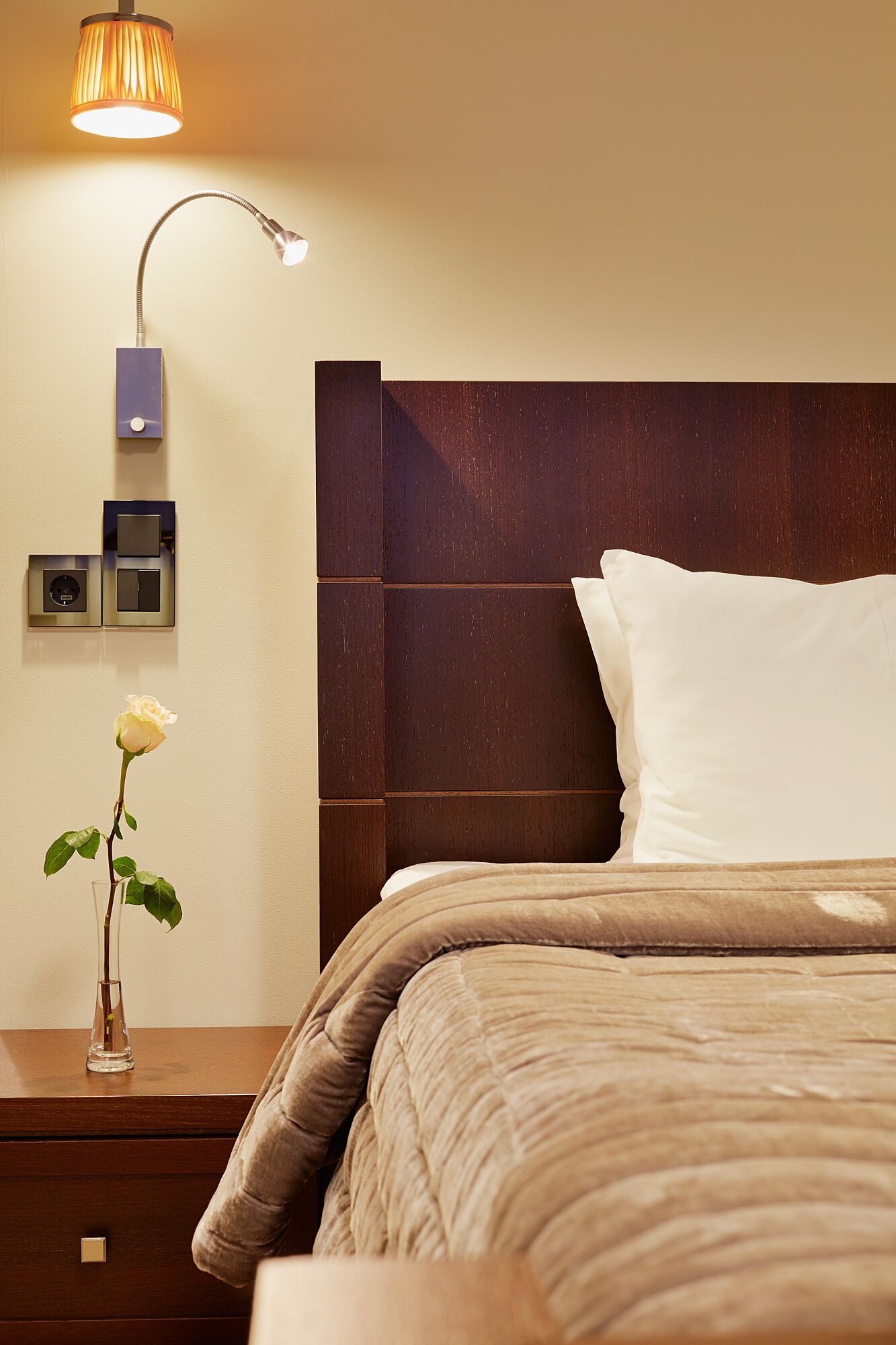 A neatly made bed with a brown headboard, a bedside lamp, a small vase with a single white rose, and a wall-mounted control panel.