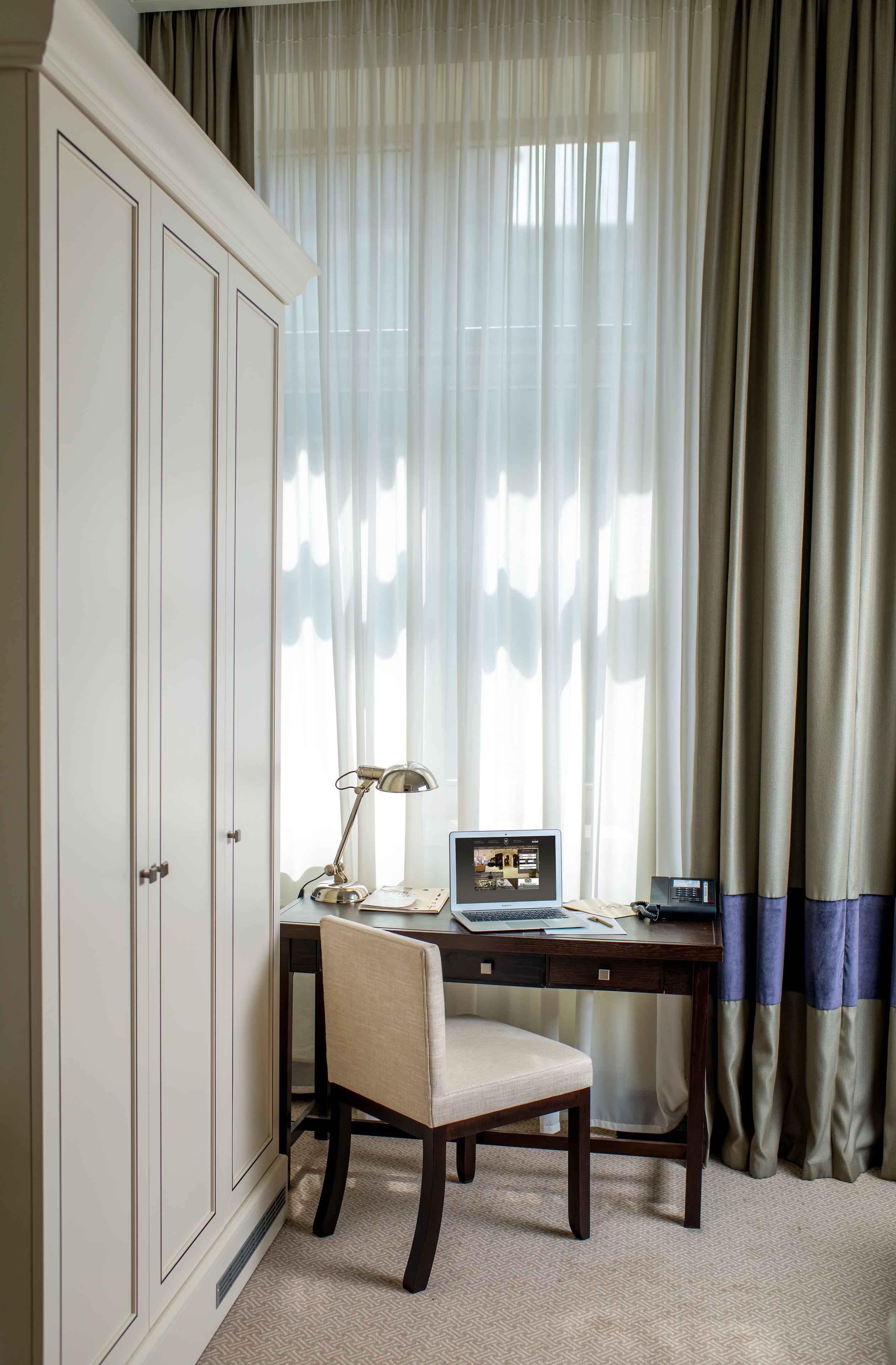 Een kamer met een bureau, stoel en een computer. Er is een raam met gordijnen en uitzicht op het omliggende landschap.