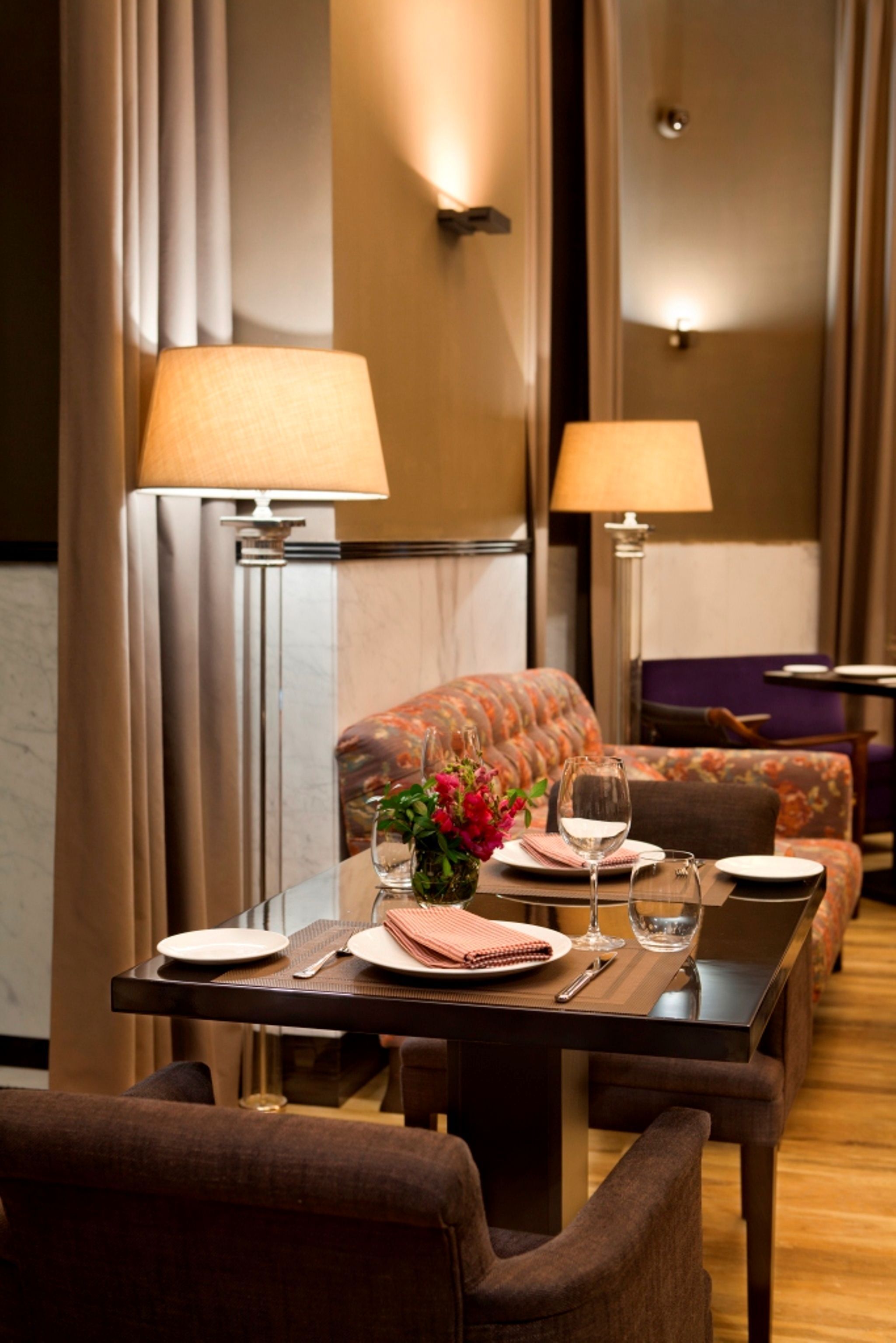 A dining area with a table set for two, featuring a vase of flowers, wine glasses, and neatly arranged plates and cutlery.