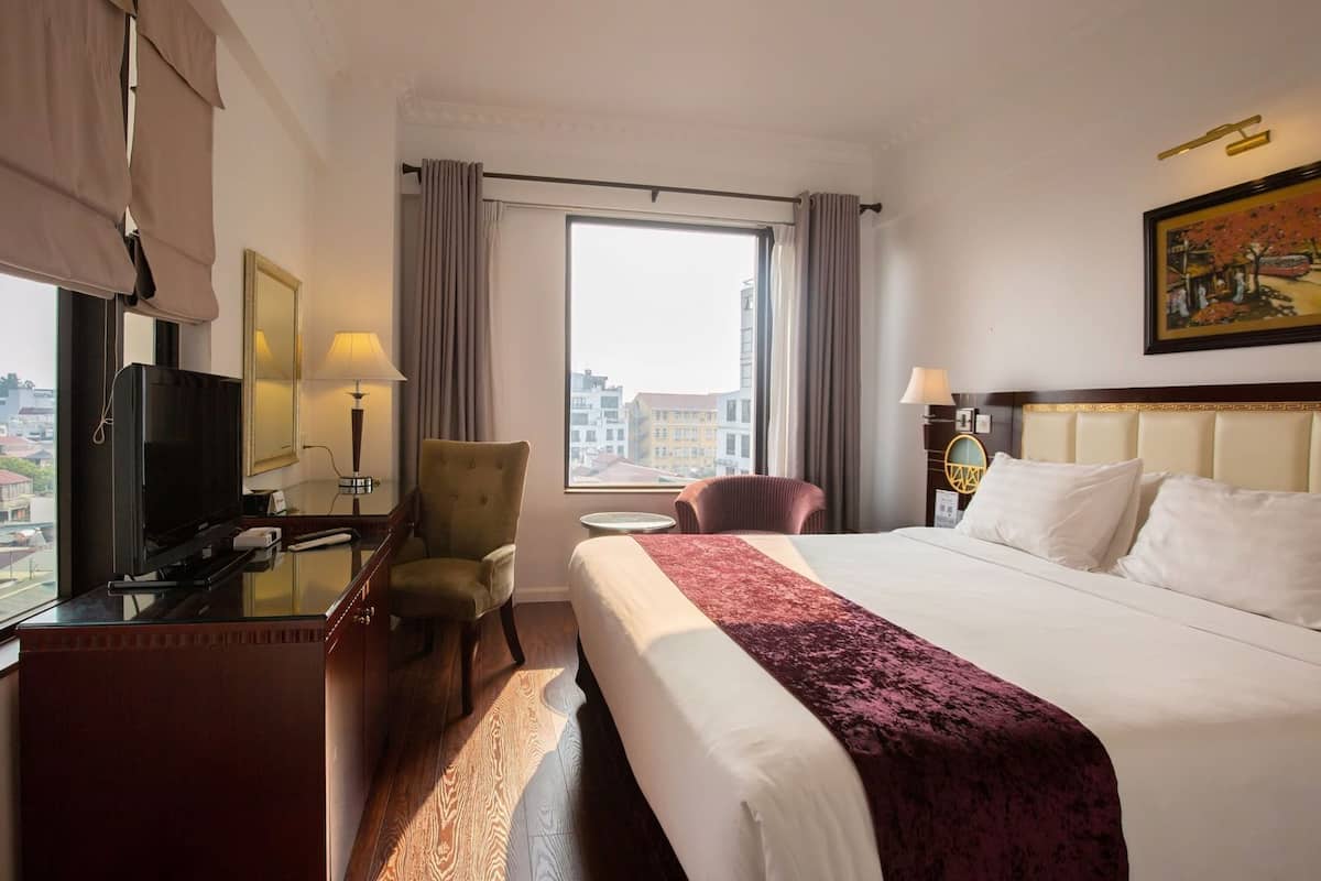 A hotel room with a bed, a desk with a TV, a chair, a window with curtains, and a painting on the wall.