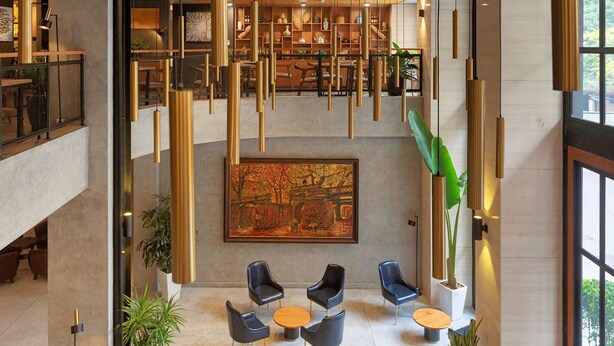 A modern lobby with a high ceiling, hanging pendant lights, a large mural, and seating area with tables and chairs. - Flower Garden Hotel (Hanoi)