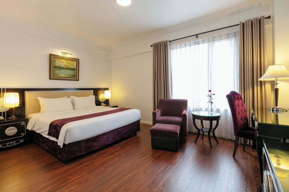 A hotel room with a large bed, a chair, a small table, and a lamp.