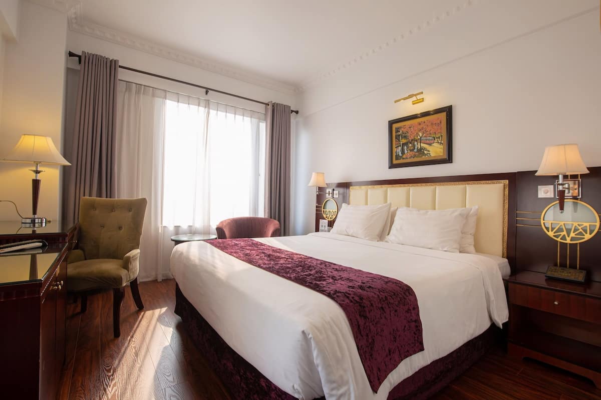 A hotel room with a large bed, a desk with a lamp, a chair, and a painting on the wall.