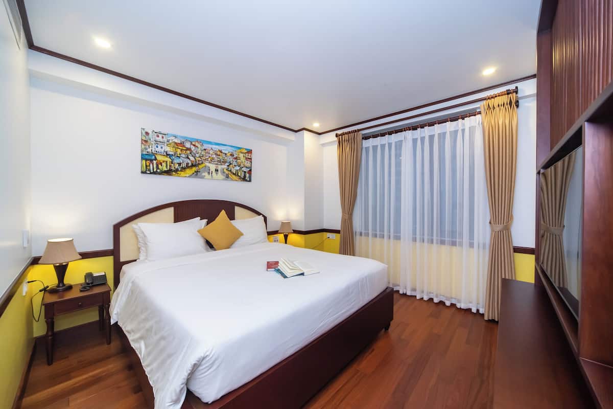 A hotel room with a large bed, wooden headboard, two bedside tables with lamps, a painting on the wall, and a window with sheer curtains.