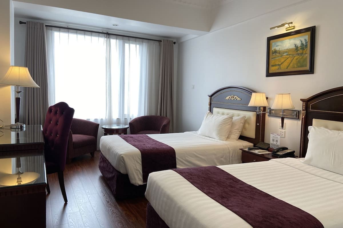 A hotel room with two beds, a desk with a lamp, a chair, a painting on the wall, and a window with curtains.