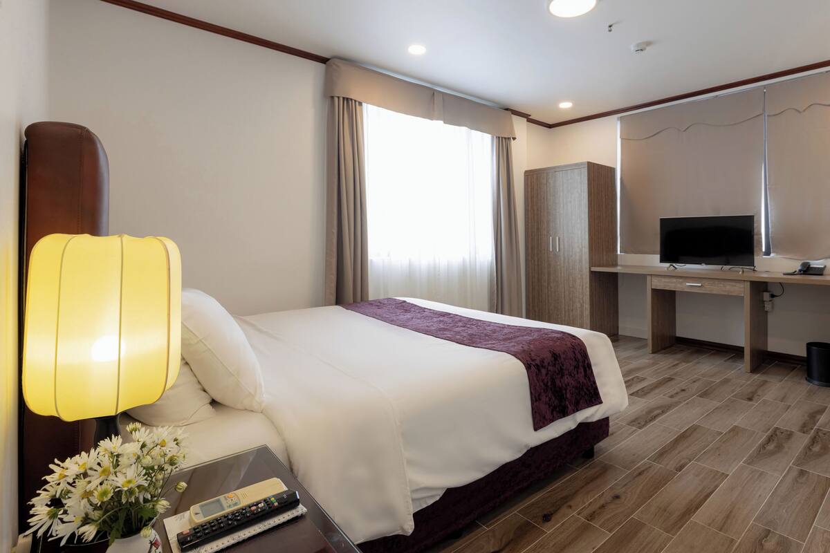 A hotel room with a bed, bedside lamp, desk with a television, and a window with curtains.