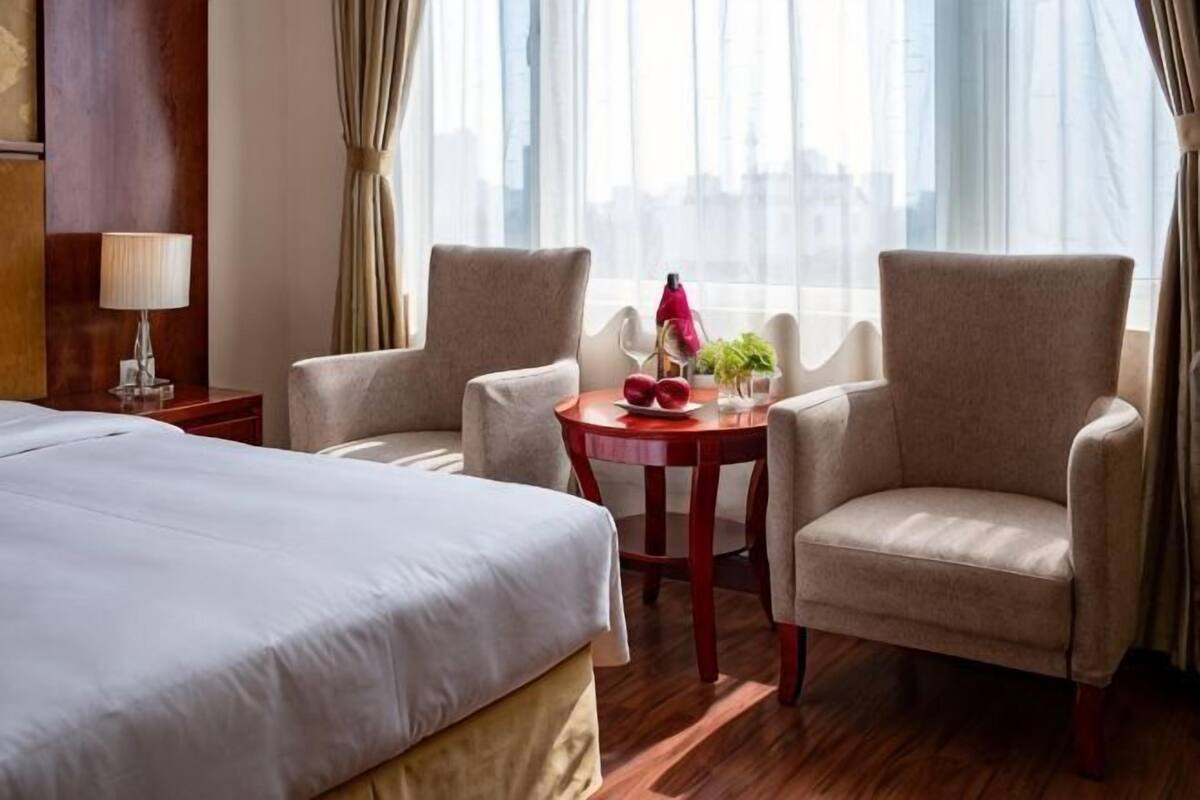 A hotel room with a bed, two armchairs, a small round table with a red object, and a lamp.