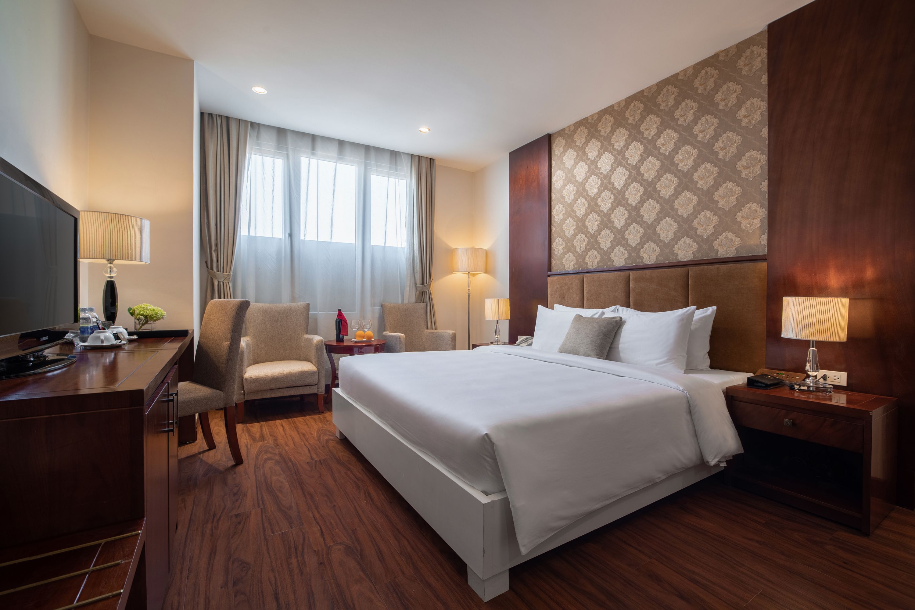 A hotel room with a large bed, a desk with a television, a chair, and a nightstand with a lamp.
