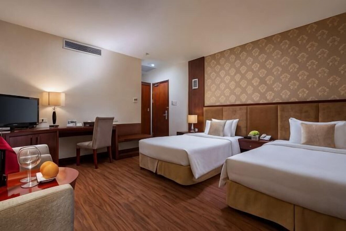 A hotel room with two beds, a desk, a chair, a television, and a small table with a fruit bowl.