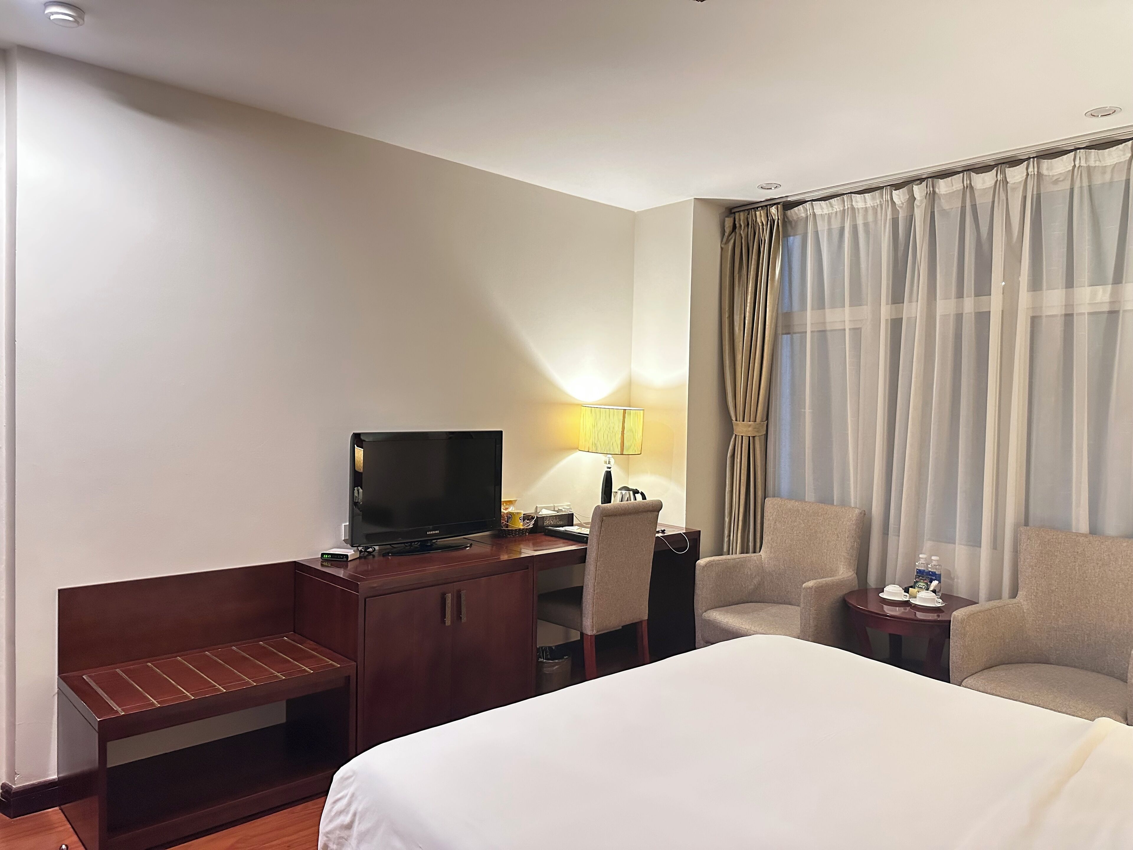 A hotel room with a bed, a TV on a wooden desk, a lamp, two chairs, and a window with curtains.