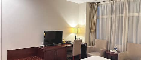 A hotel room with a bed, a TV on a wooden desk, a lamp, two chairs, and a window with curtains.
