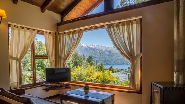 A cozy living room with a sofa, a TV, a table, and a fireplace. Large windows offer a scenic view of mountains and a lake. - Estancia del Carmen (San Carlos de Bariloche)