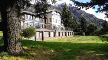 A two-story building with a balcony, surrounded by a grassy area and trees, with mountains in the background.