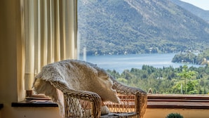 A wicker chair with a white, fluffy throw blanket, positioned by a window overlooking a mountainous landscape and a lake. - Estancia del Carmen (San Carlos de Bariloche)