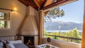 A cozy room with a fireplace, a sofa, and a table with a decorative item. A window offers a view of mountains and a lake. - Estancia del Carmen (San Carlos de Bariloche)