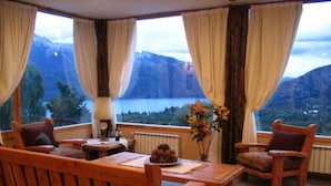 A wooden-furnished room with a table and chairs, large windows with curtains, a view of mountains and a lake, and a vase with flowers on the table. - Estancia del Carmen (San Carlos de Bariloche)