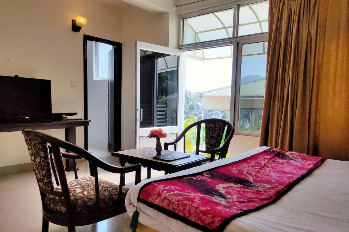 Ein Hotelzimmer mit Bett, Schreibtisch, zwei Stühlen, Fernseher und einem Fenster mit Blick auf Grünflächen.
