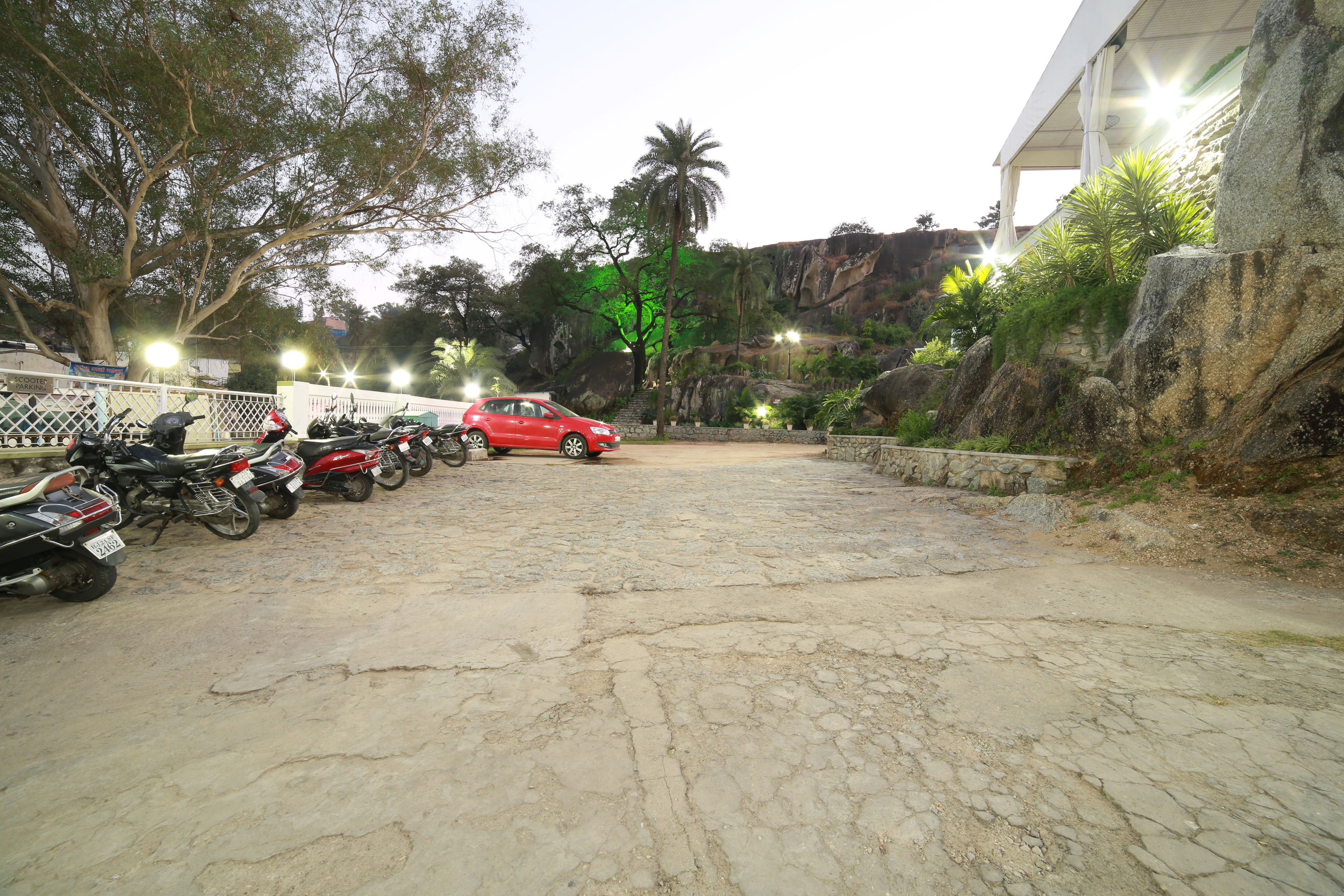 Un estacionamiento con motocicletas y un coche rojo, rodeado de árboles y un terraplén rocoso.