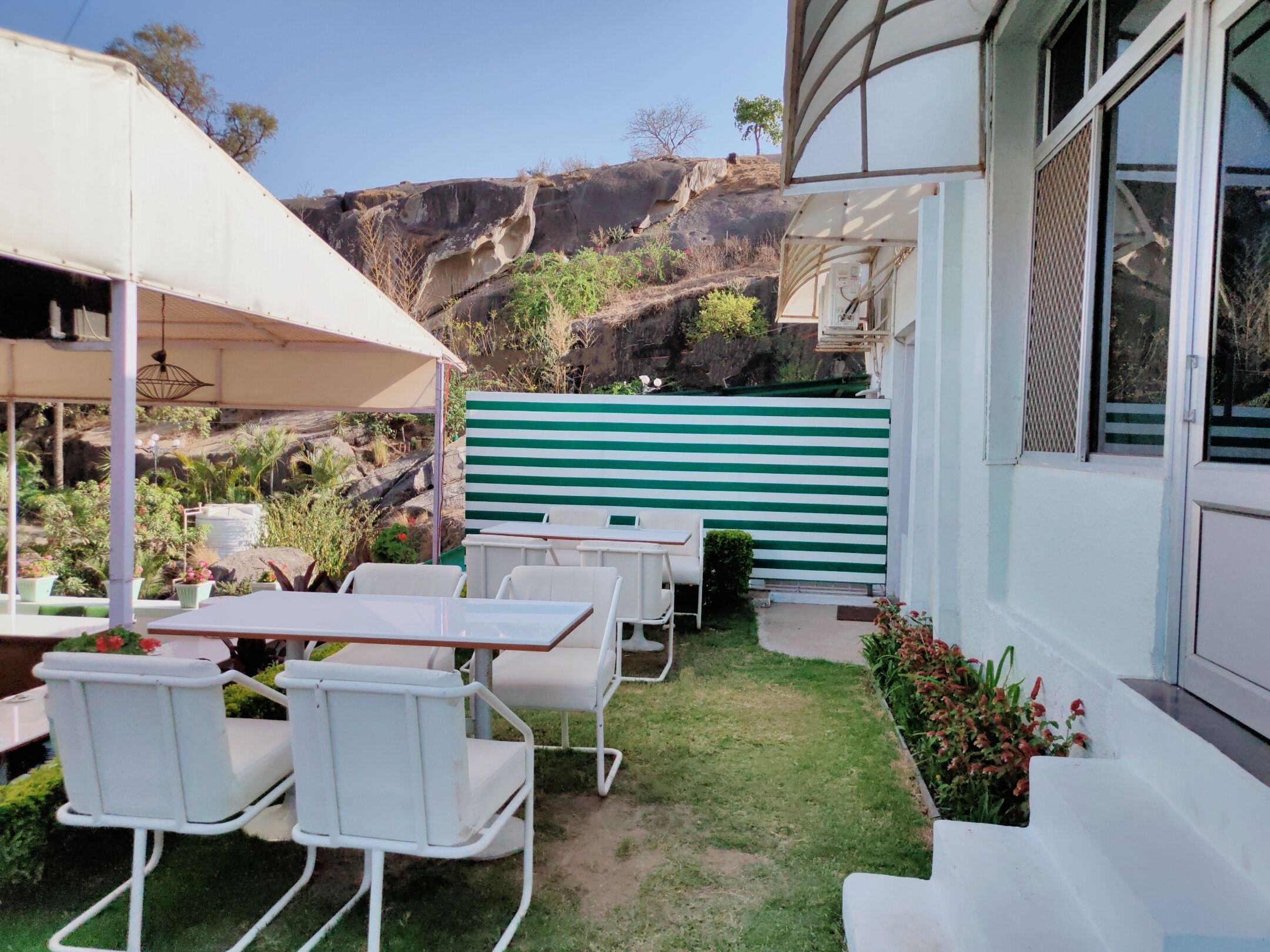 An outdoor patio with white metal chairs and a table, a green and white striped fence, and a view of rocky hills.