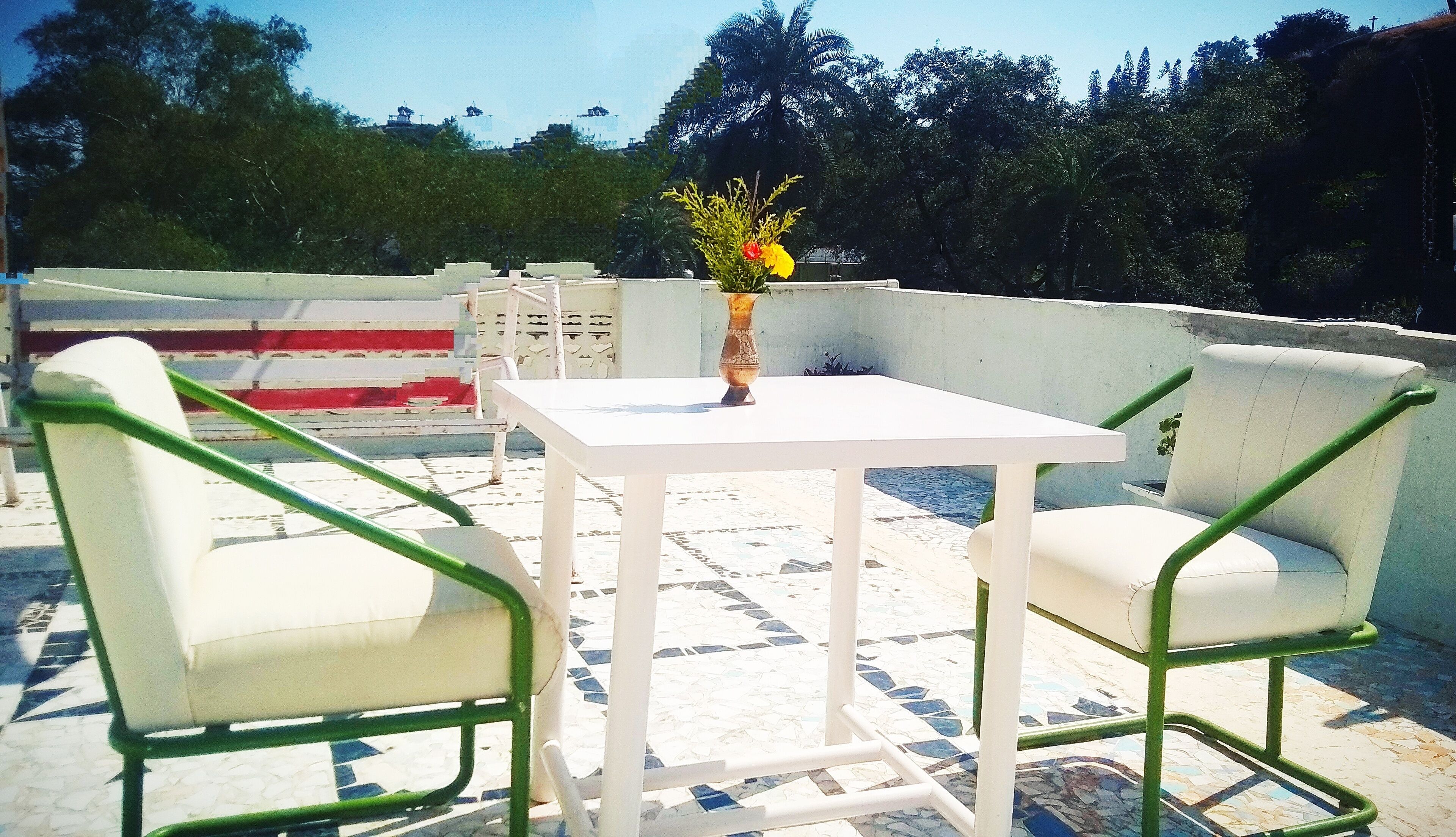 A terrace with a white table and two white chairs with green frames, a vase with flowers, and a tiled floor.