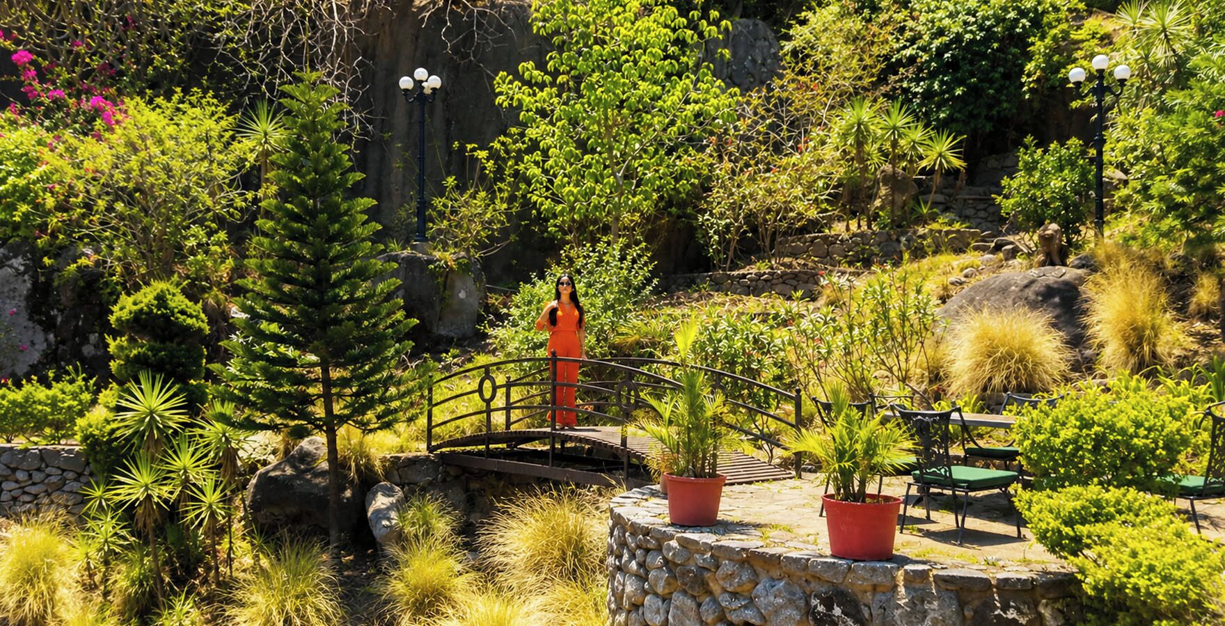 Un jardin avec un mur en pierre, des plantes en pot, un pont en bois et du mobilier d’extérieur.